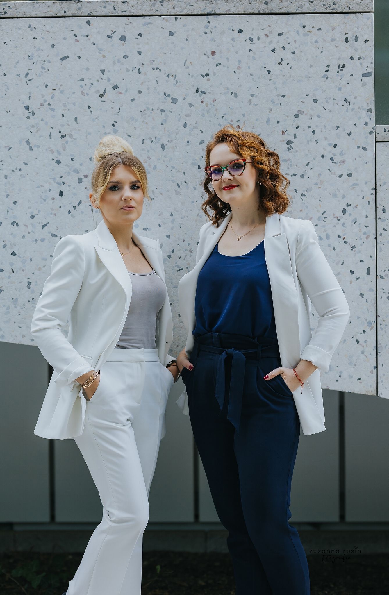 Dwie kobiety w eleganckich strojach stojące na tle ściany z mozaiką, blondynka z włosami upiętymi w kok, brunetka w okularach z czerwoną oprawką.