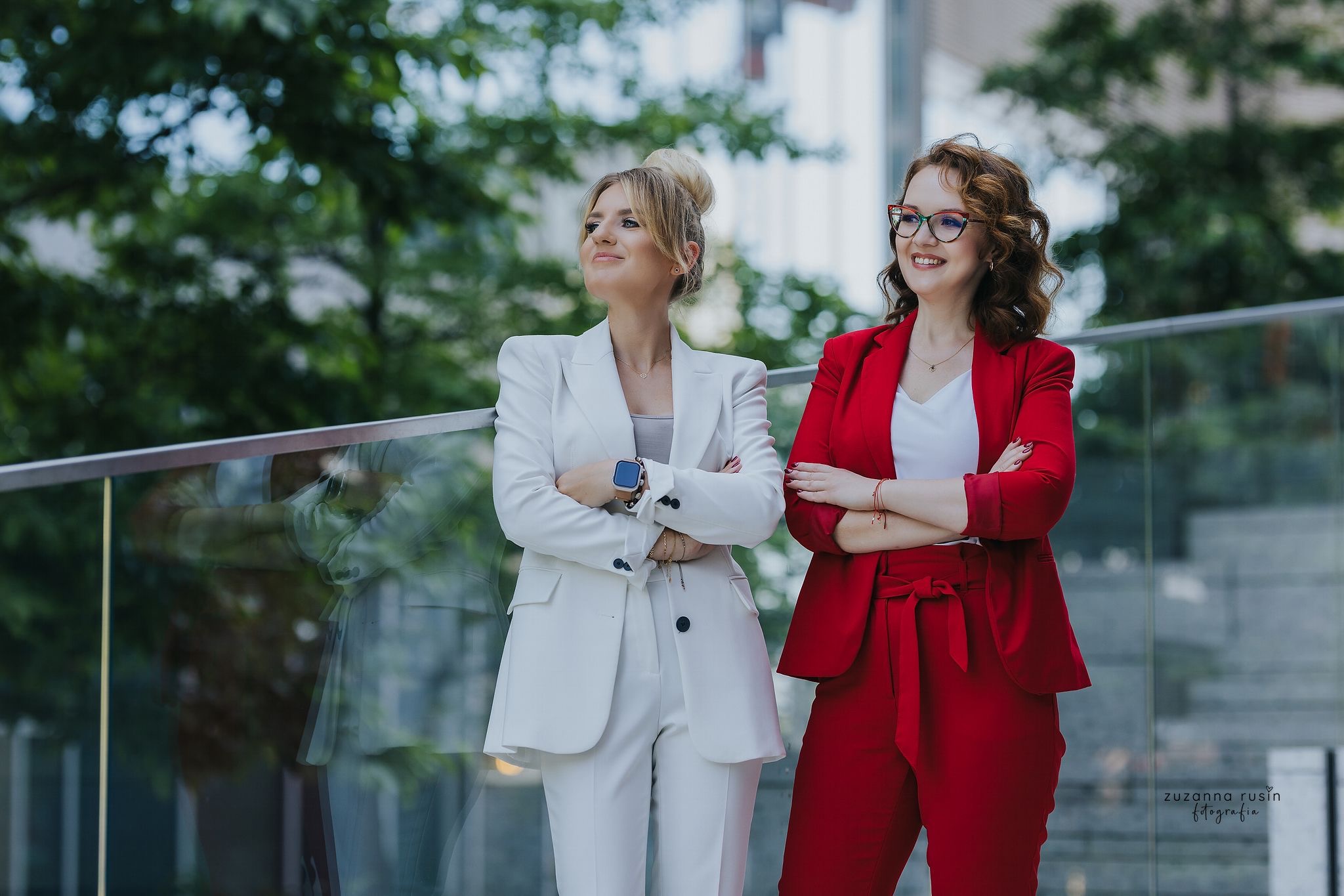Dwie uśmiechnięte kobiety w eleganckich garniturach, białym i czerwonym, stoją obok siebie w Warszawie na tle zieleni i nowoczesnej architektury.
