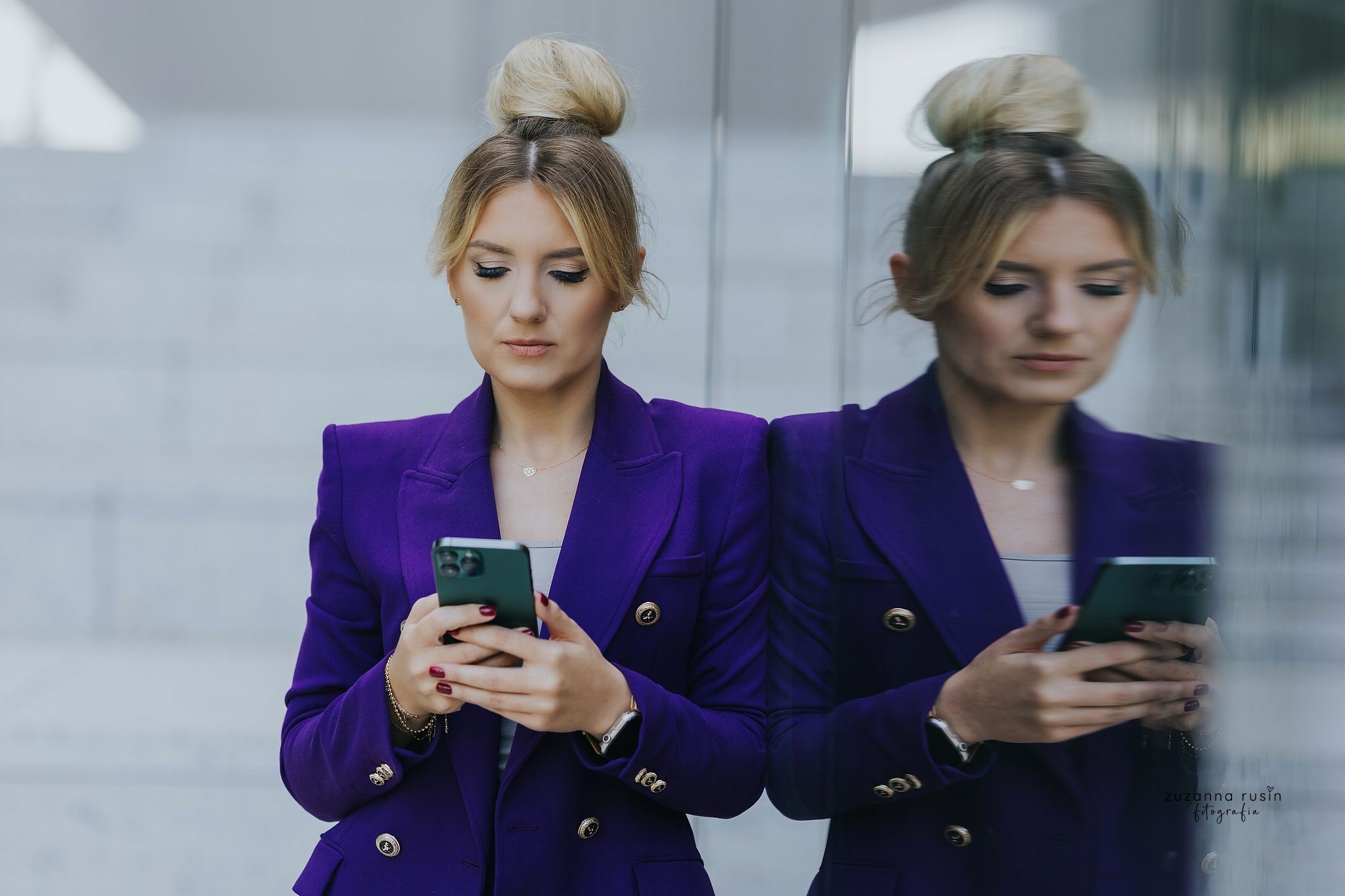 Kobieta w fioletowym żakiecie, z upiętymi blond włosami, sprawdza coś w telefonie, odbijając się w szklanej powierzchni.