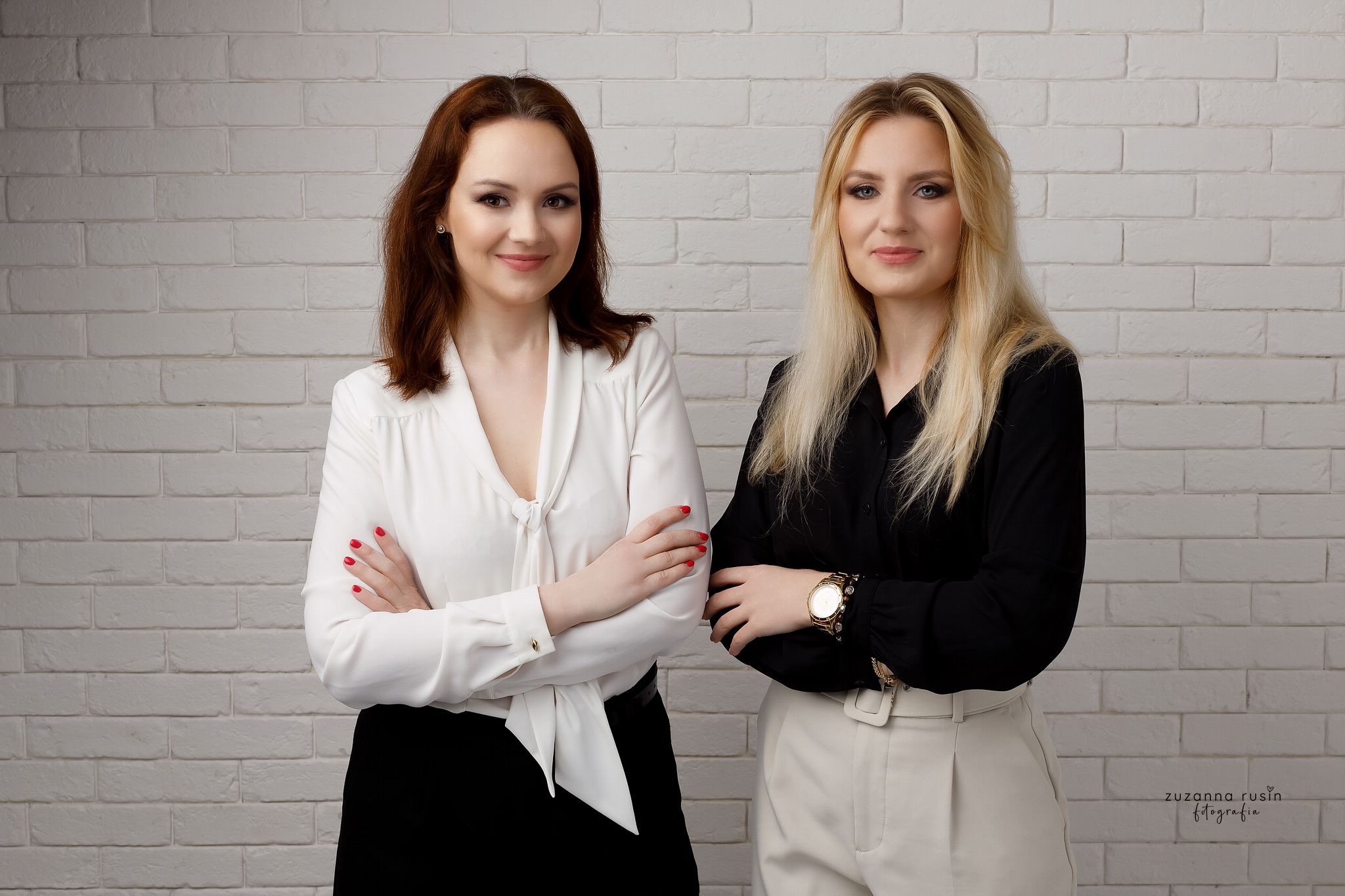 Portret dwóch uśmiechniętych kobiet w eleganckich strojach na tle ściany z białej cegły, jedna w białej bluzce i czarnej spódnicy, druga w czarnej bluzce i beżowych spodniach.