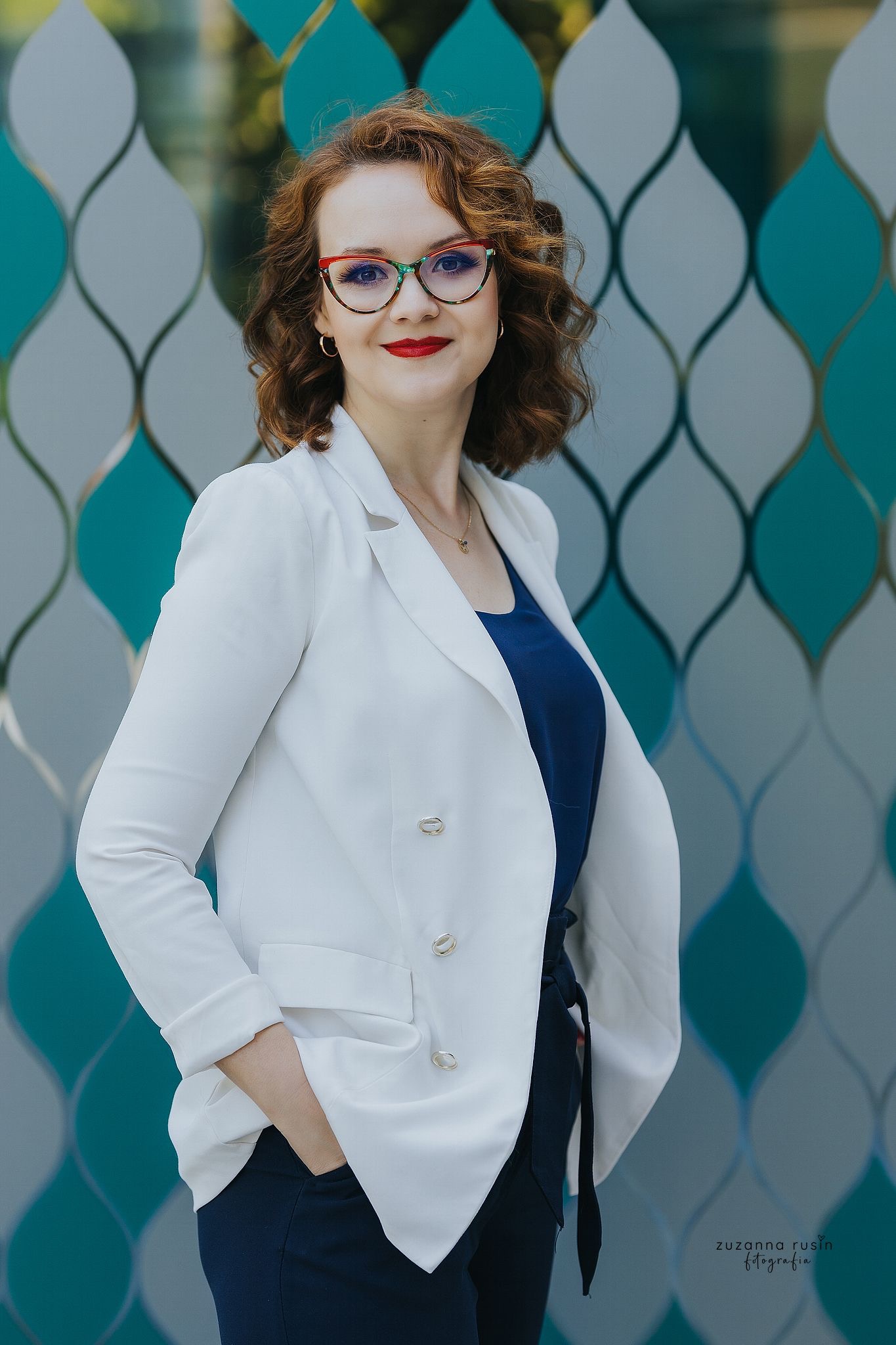 Uśmiechnięta kobieta z rudymi włosami w okularach w ozdobnych oprawkach, ubrana w białą marynarkę i granatową bluzkę, pozuje na tle dekoracyjnej ściany w odcieniach błękitu i szarości.