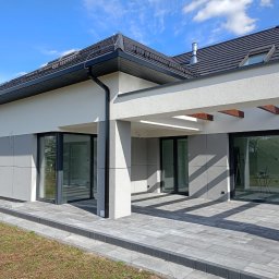 WKBUD - Nowoczesny dom jednorodzinny z szarą elewacją, dużymi przesuwnymi oknami i zadaszonym tarasem z drewnianymi belkami, widok z zewnątrz na wybrukowany taras i fragment trawnika.