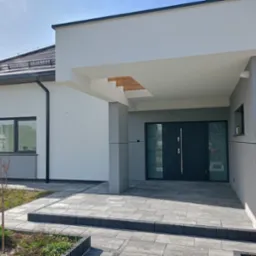 Nowoczesny dom jednorodzinny z szarą elewacją, widoczne wejście z zadaszeniem i drewnianym elementem dekoracyjnym, grafitowe drzwi wejściowe i okna, kostka brukowa na podjeździe.