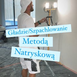 WKBUD - Mężczyzna w białym ubraniu natryskuje gładź na ścianę za pomocą specjalnego agregatu, oświetlony dwoma halogenami na statywie, podłoga zabezpieczona folią.