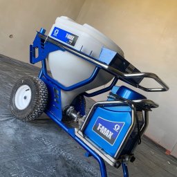 WKBUD - Profesjonalne urządzenie malarskie Graco T-Max 506 na kółkach, z białym zbiornikiem i niebieską ramą, ustawione na czarnej folii ochronnej w pomieszczeniu przed malowaniem ścian.