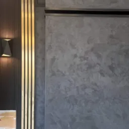 Dekoracyjna ściana z efektem betonu architektonicznego, sąsiadująca z panelem ściennym w kolorze grafitowym z pionowymi, złotymi listwami i nowoczesnym kinkietem.