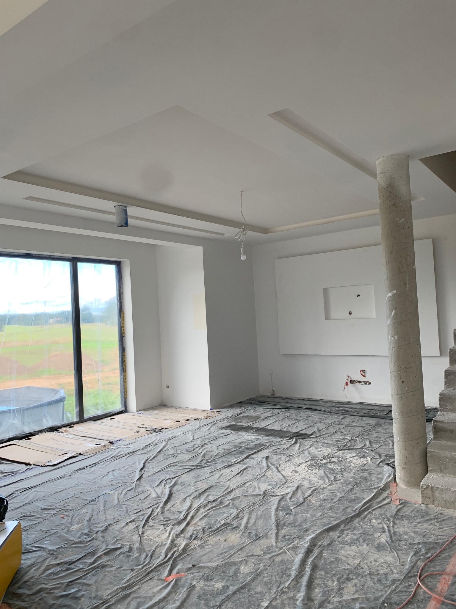 Wnętrze w trakcie prac wykończeniowych: zabezpieczona folią podłoga, surowy betonowy filar, ściany przygotowane pod malowanie, widoczne elementy instalacji elektrycznej.