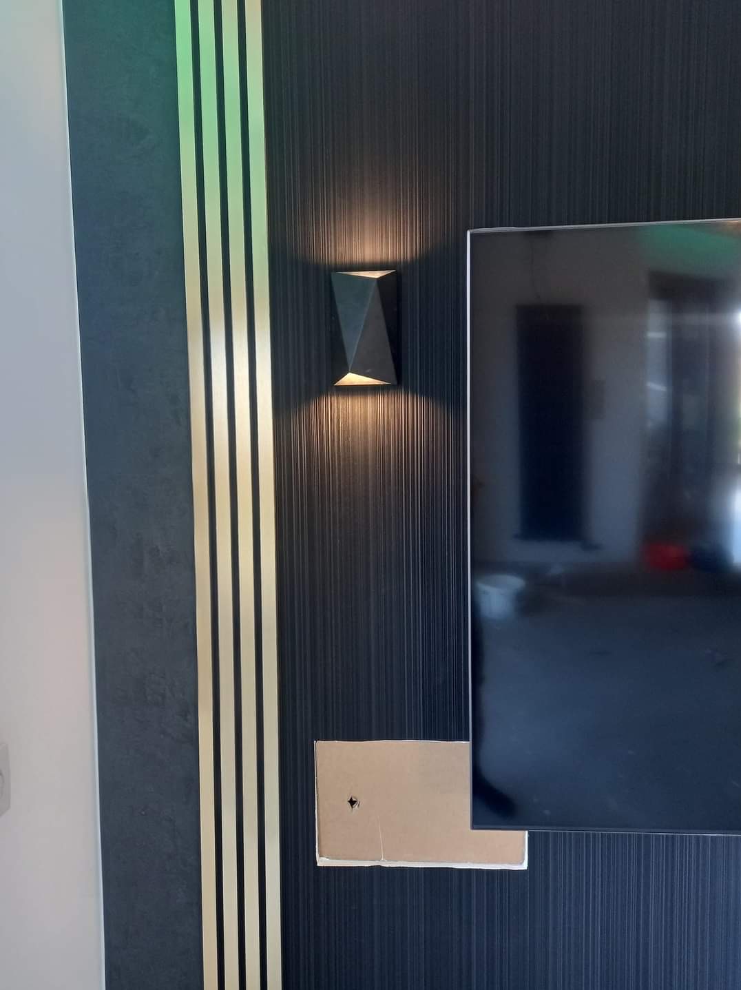 Ciemna ściana z pionowymi paskami w odcieniach szarości i złota, ozdobiona geometrycznym kinkietem i fragmentem telewizora z widocznym otworem montażowym.