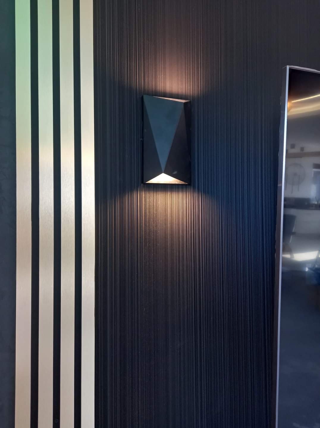 Czarna, geometryczna lampa ścienna oświetla pionową, teksturowaną ścianę w odcieniach granatu, sąsiadującą z dekoracyjnymi, złotymi listwami.