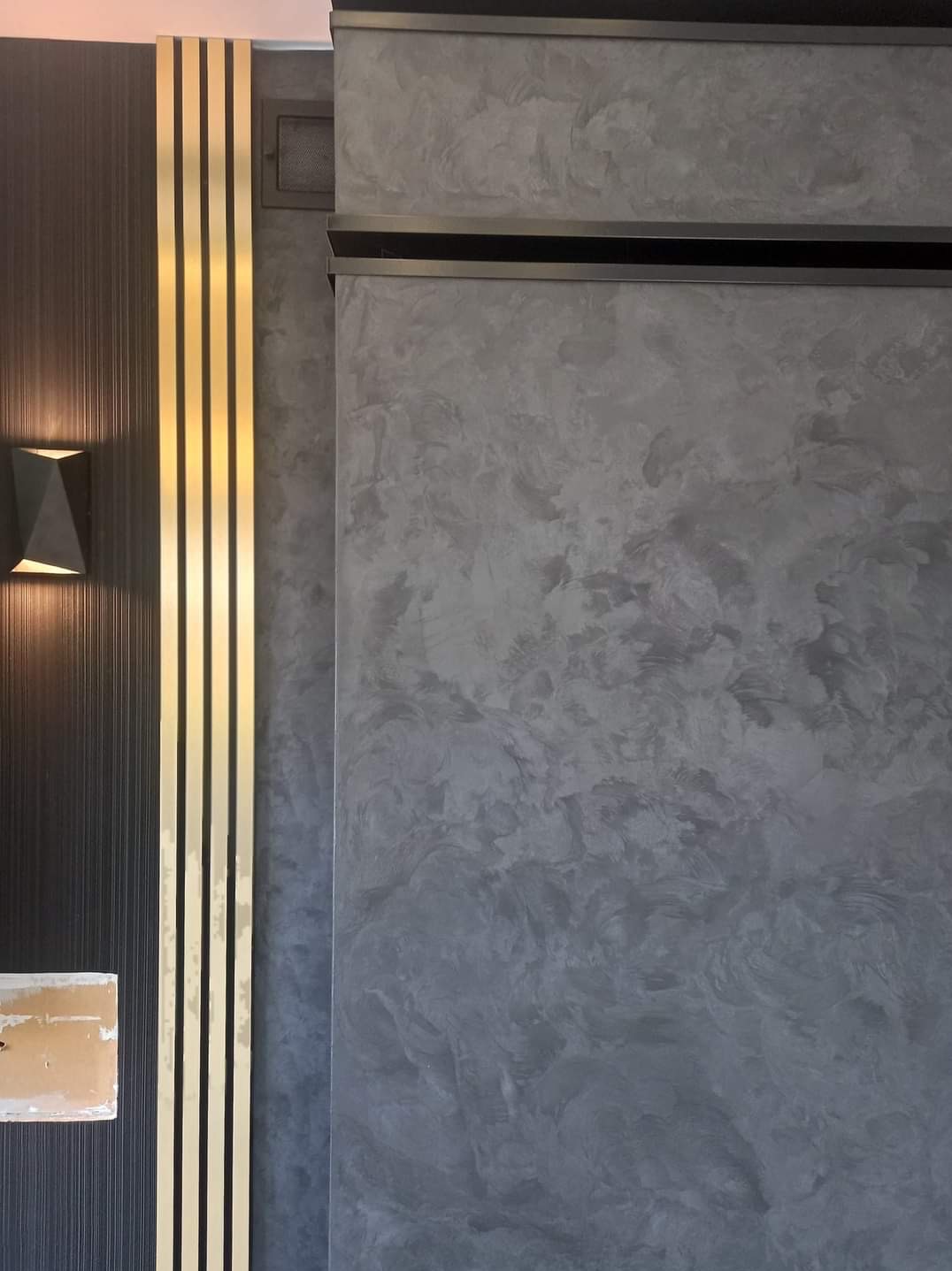 Dekoracyjna ściana z efektem betonu architektonicznego, sąsiadująca z panelem ściennym w kolorze grafitowym z pionowymi, złotymi listwami i nowoczesnym kinkietem.
