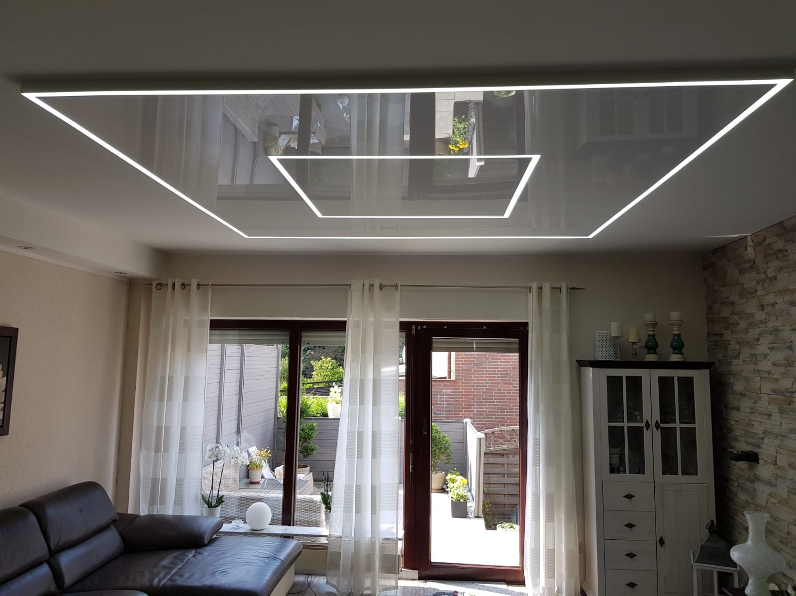 Nowoczesne oświetlenie sufitowe z podwieszanym, lustrzanym sufitem i wbudowanymi liniami LED, odbijające widok na zewnątrz przez okna z białymi zasłonami.