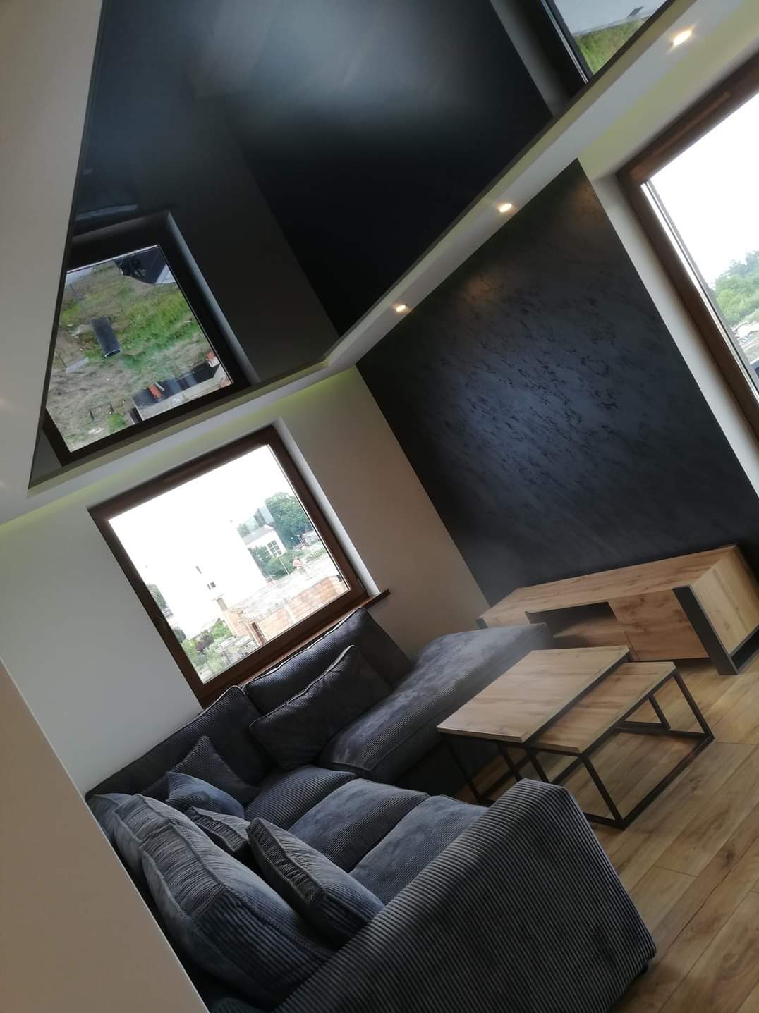 Nowoczesny salon z ciemnym, lustrzanym sufitem podwieszanym, ścianą o fakturze betonu, drewnianą podłogą i szarym narożnikiem z poduszkami.