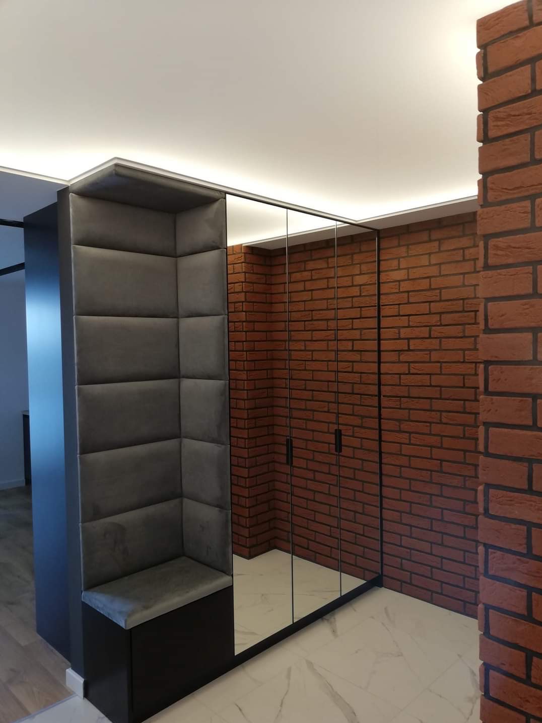 Nowoczesna aranżacja przedpokoju z tapicerowanym siedziskiem narożnym w kolorze grafitowym, lustrzaną szafą w czarnej ramie i ścianą z czerwonej cegły.