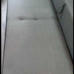 NT Detailing Profesjonalne Czyszczenie Tapicerki - Jasnoszara kanapa z dwoma wgłębieniami pośrodku siedziska, widoczny fragment boku z uchwytem i ciemnobrązowe obramowanie dolnej części.