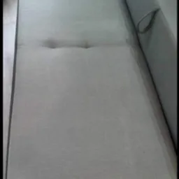 Jasnoszara kanapa z dwoma wgłębieniami pośrodku siedziska, widoczny fragment boku z uchwytem i ciemnobrązowe obramowanie dolnej części.