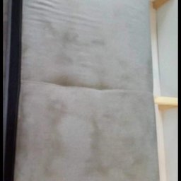 NT Detailing Profesjonalne Czyszczenie Tapicerki - Szary materac kanapy z widocznymi plamami i zabrudzeniami, ustawiony obok białego regału z drewnianymi półkami, ujęcie z góry.