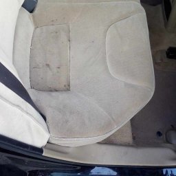 NT Detailing Profesjonalne Czyszczenie Tapicerki - Brudna, beżowa tapicerka tylnej kanapy samochodu, widoczne plamy i zacieki, częściowo otwarte drzwi auta, kostka brukowa na zewnątrz.