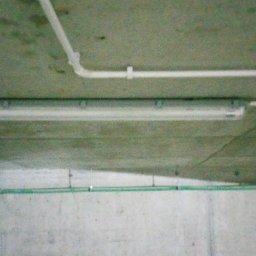 NT Detailing Profesjonalne Czyszczenie Tapicerki - Zamontowana pod sufitem betonowym lampa fluorescencyjna z widocznymi przewodami elektrycznymi w białych plastikowych rurkach i zielony przewód w dolnej części ściany.