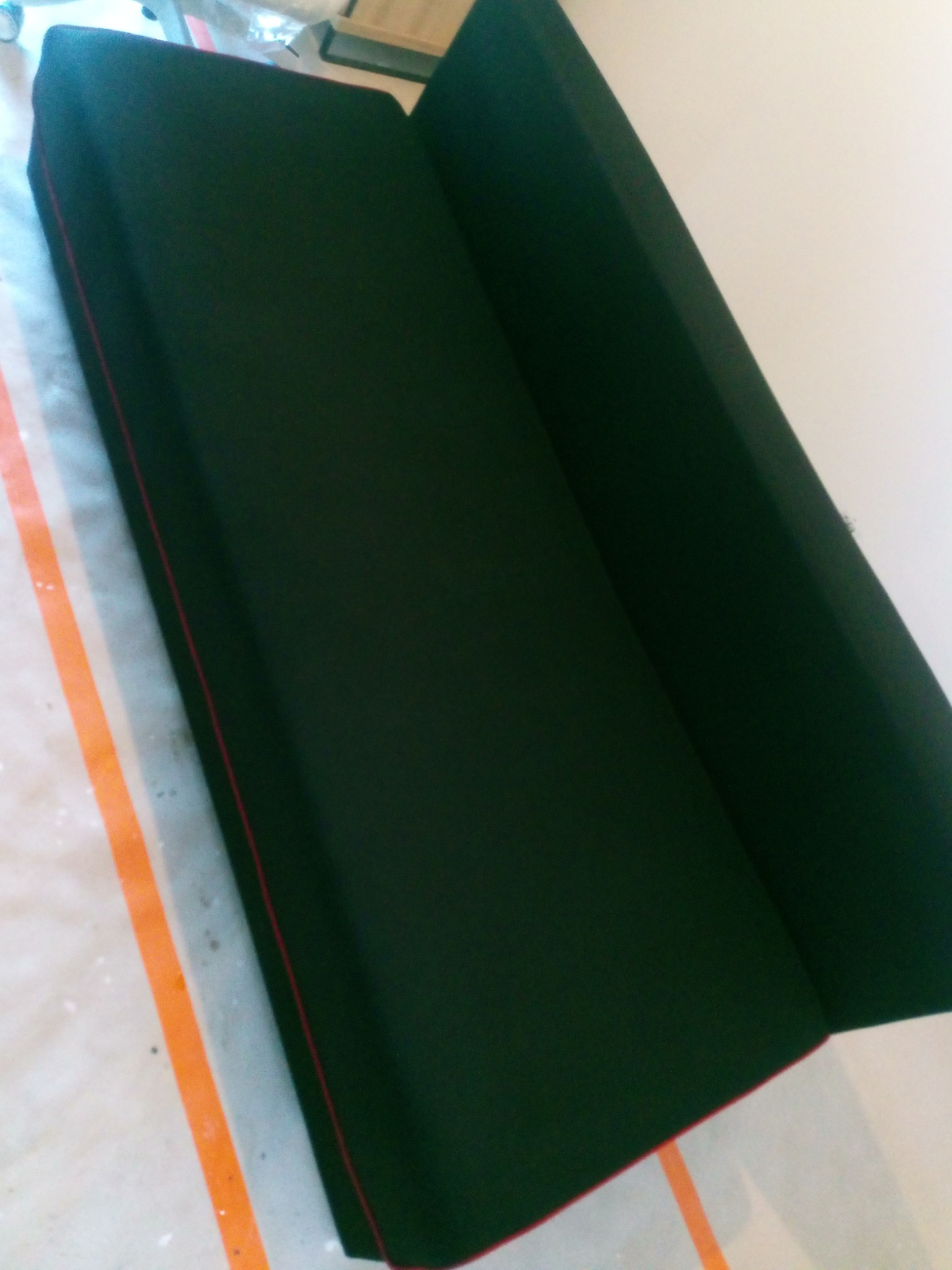 Ciemnozielona kanapa z czerwoną lamówką stoi na zabezpieczonej folią podłodze, gotowa do użytku po czyszczeniu.