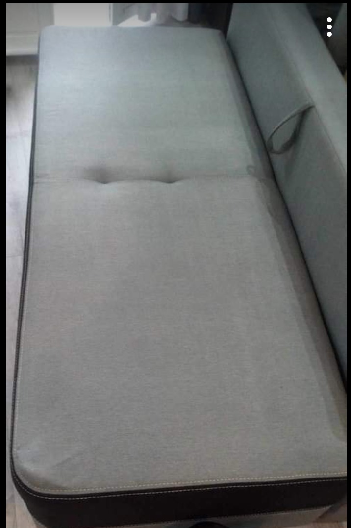 Jasnoszara kanapa z dwoma wgłębieniami pośrodku siedziska, widoczny fragment boku z uchwytem i ciemnobrązowe obramowanie dolnej części.