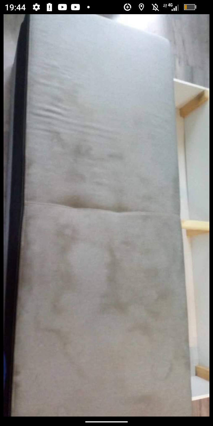 Szary materac kanapy z widocznymi plamami i zabrudzeniami, ustawiony obok białego regału z drewnianymi półkami, ujęcie z góry.