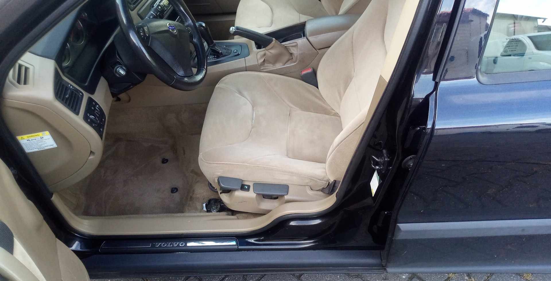 Wnętrze samochodu Volvo z beżową tapicerką przednich foteli i dywaników, widoczne zabrudzenia na siedzeniu kierowcy, otwarte drzwi.