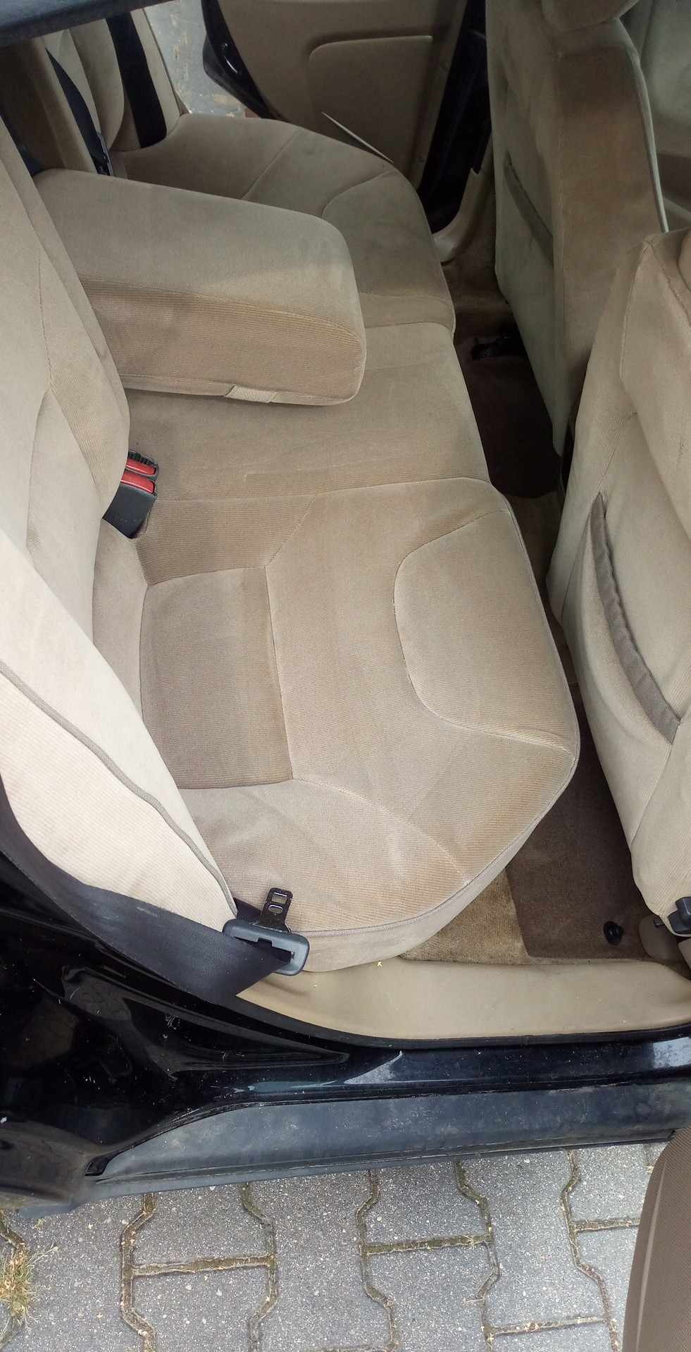 Beżowa, welurowa tapicerka tylnej kanapy samochodu z widocznymi śladami użytkowania i niewielkimi zabrudzeniami, z podłokietnikiem i pasami bezpieczeństwa.