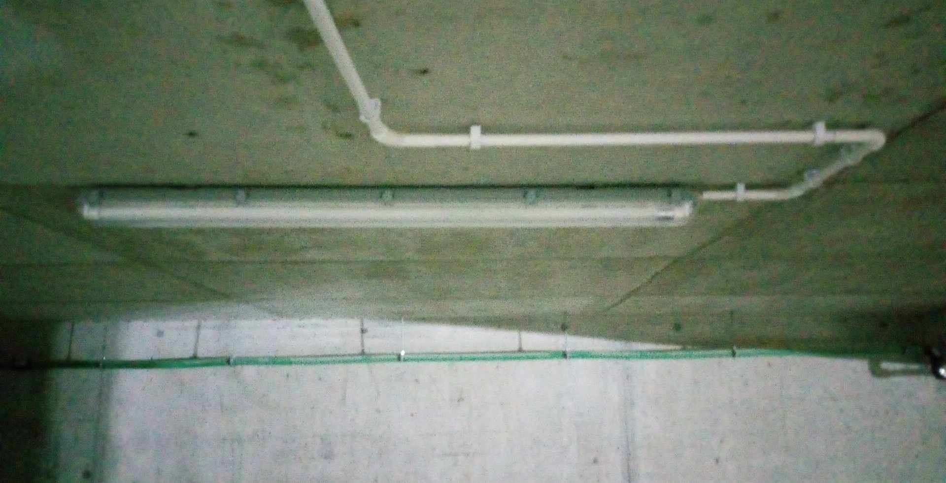 Zamontowana pod sufitem betonowym lampa fluorescencyjna z widocznymi przewodami elektrycznymi w białych plastikowych rurkach i zielony przewód w dolnej części ściany.