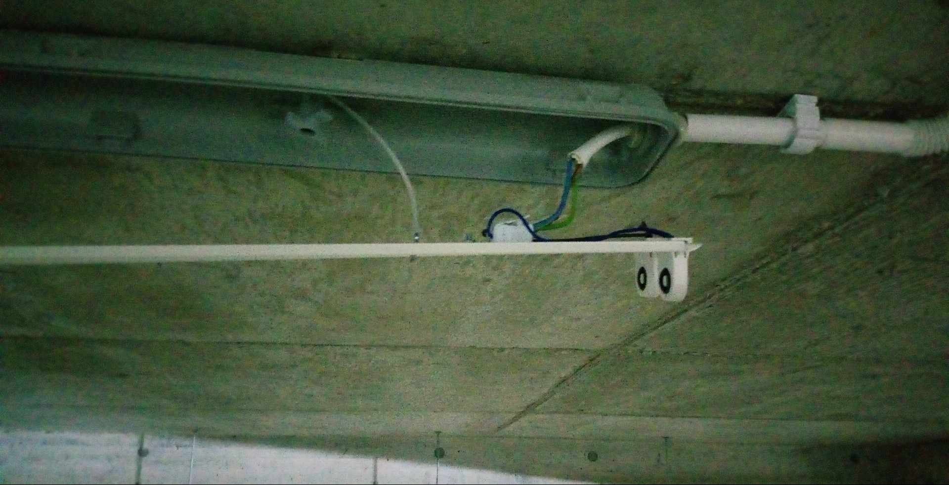 Widok z dołu na zamontowaną lampę LED z widocznymi przewodami, zamocowaną do sufitu z betonu architektonicznego, z widoczną rurą ochronną na przewody.