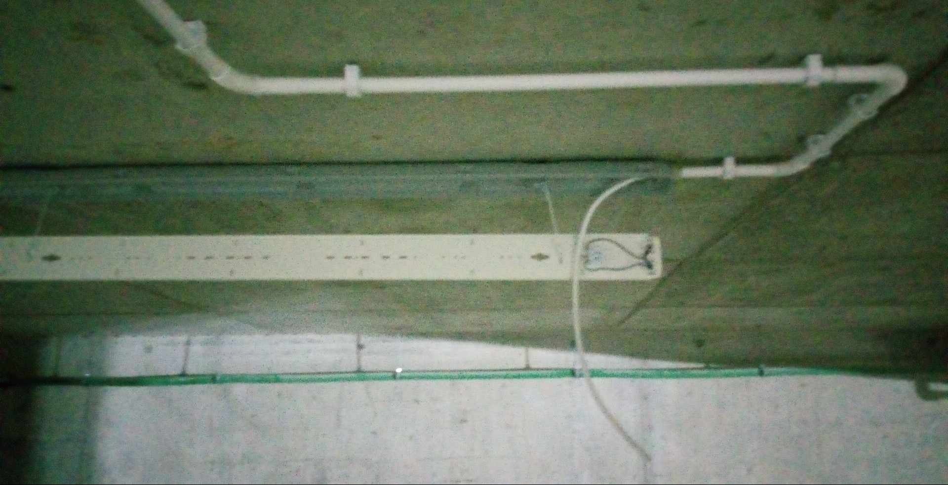 Niedokończona instalacja elektryczna na suficie z widocznymi kablami, rurami PCV i listwą oświetleniową. Zdjęcie zrobione pod kątem, w słabym oświetleniu.