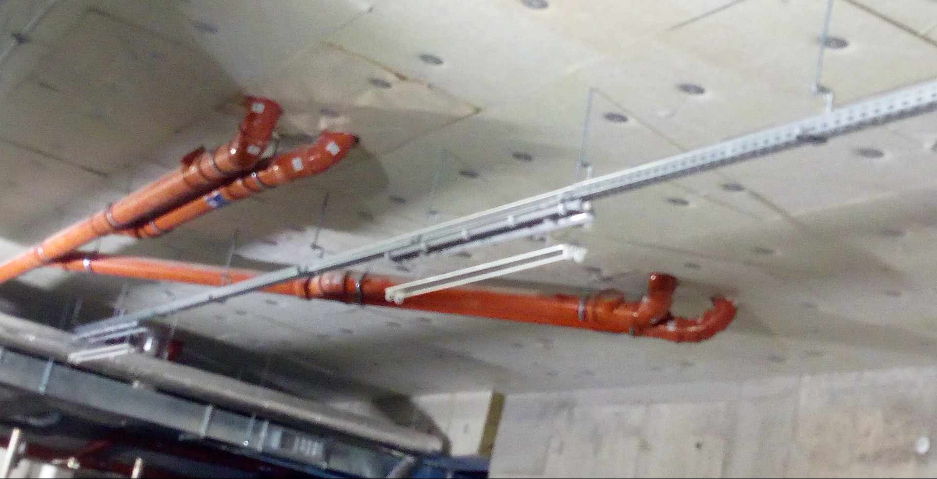 Pomarańczowe rury kanalizacyjne zamontowane pod betonowym sufitem, widoczne metalowe szyny i uchwyty mocujące.