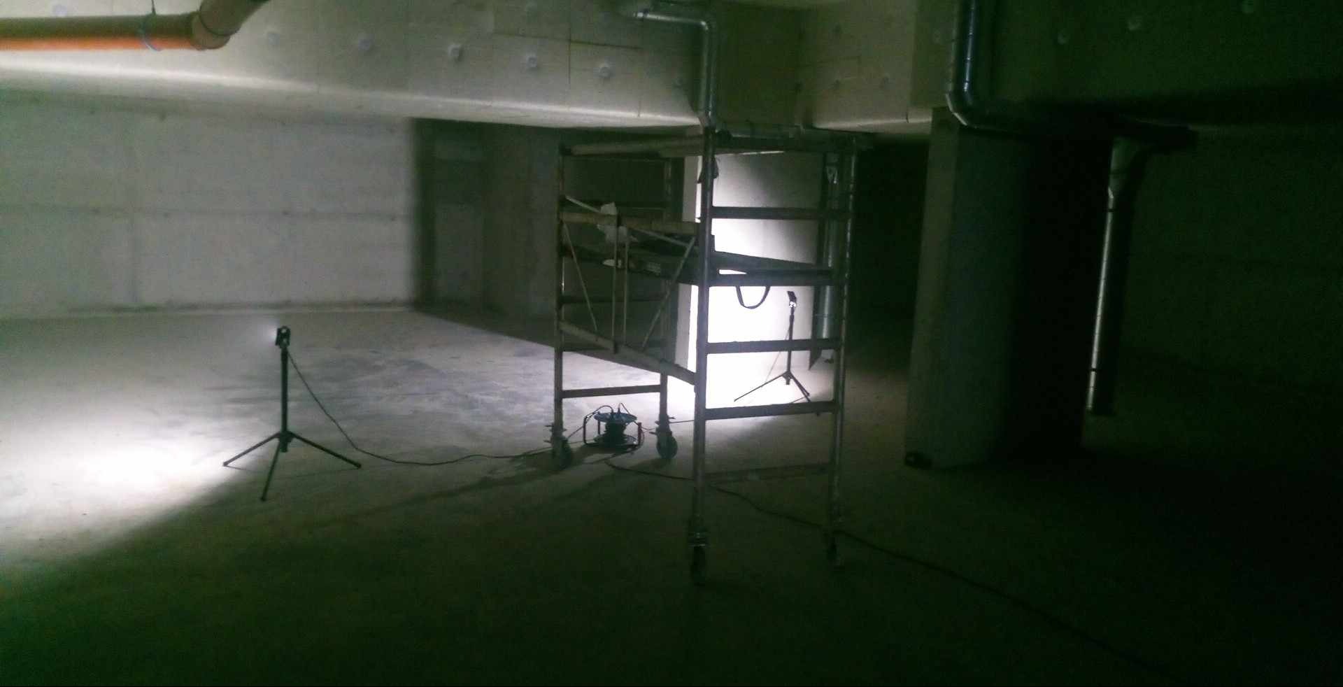 Wnętrze w surowym stanie z widocznymi rurami pod sufitem, oświetlone lampą na statywie i dodatkowo przez reflektor na rusztowaniu.
