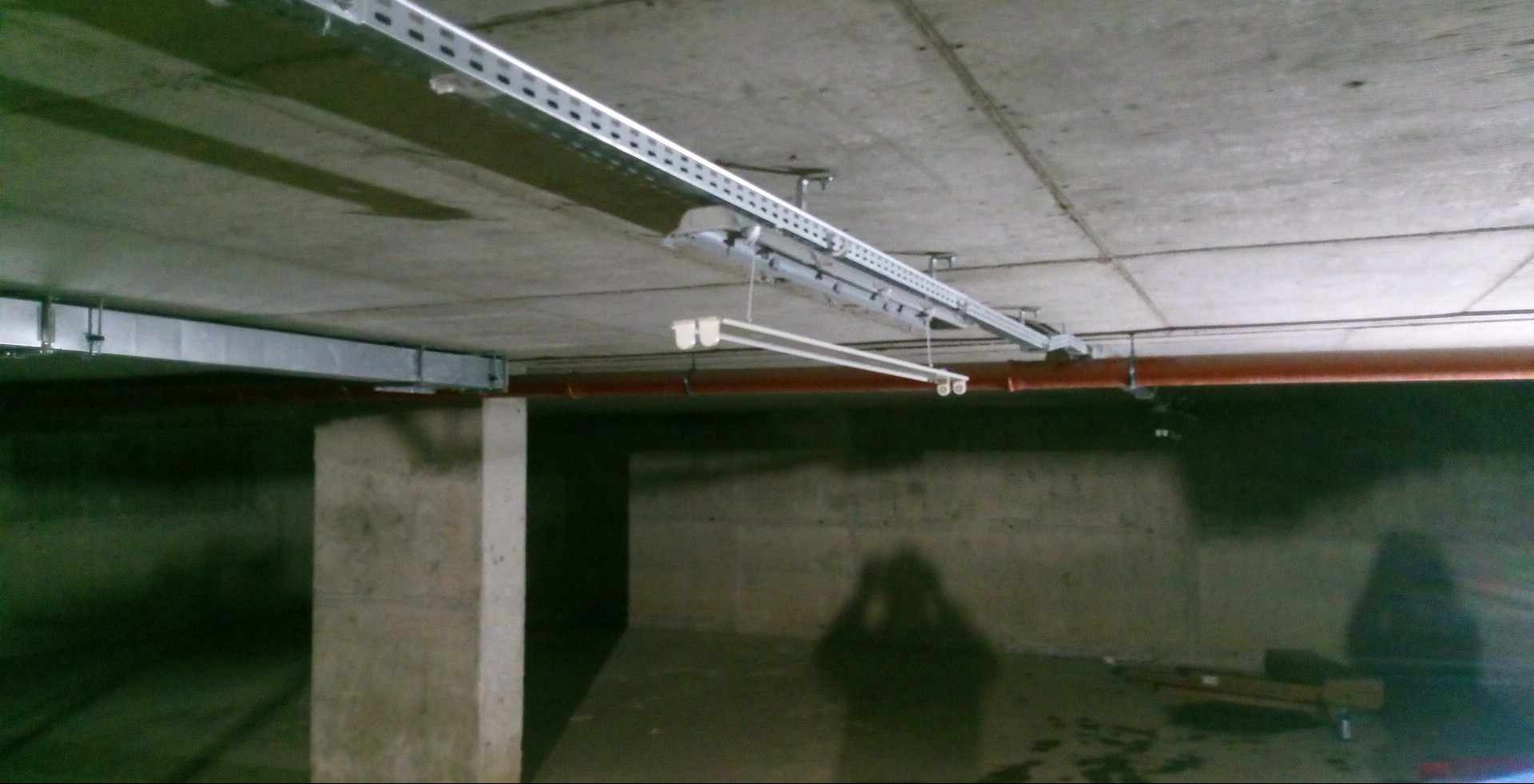 Betonowy strop z widocznymi instalacjami: koryta kablowe, rury w kolorze ceglastym i zawieszona lampa jarzeniowa. Cień osoby fotografującej na ścianie.