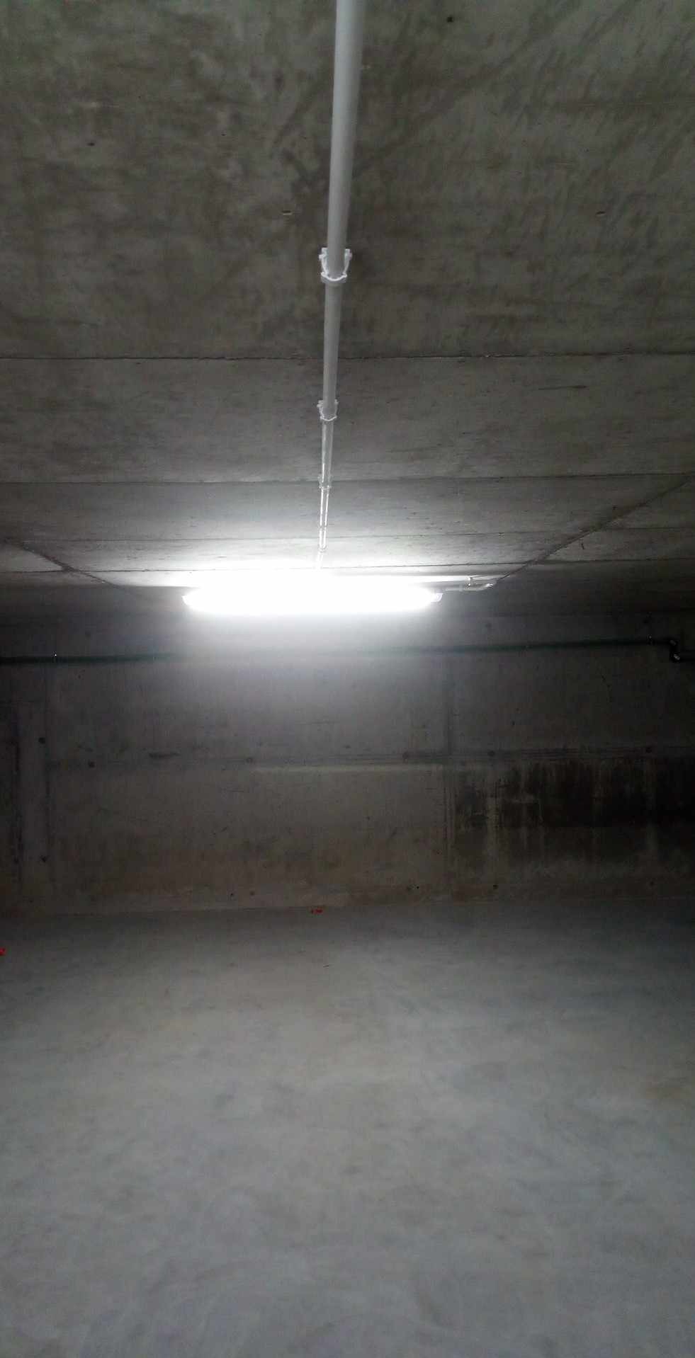 Surowe wnętrze z betonowym sufitem, widoczne rury i oświetlenie jarzeniowe, surowy beton na ścianach i podłodze.