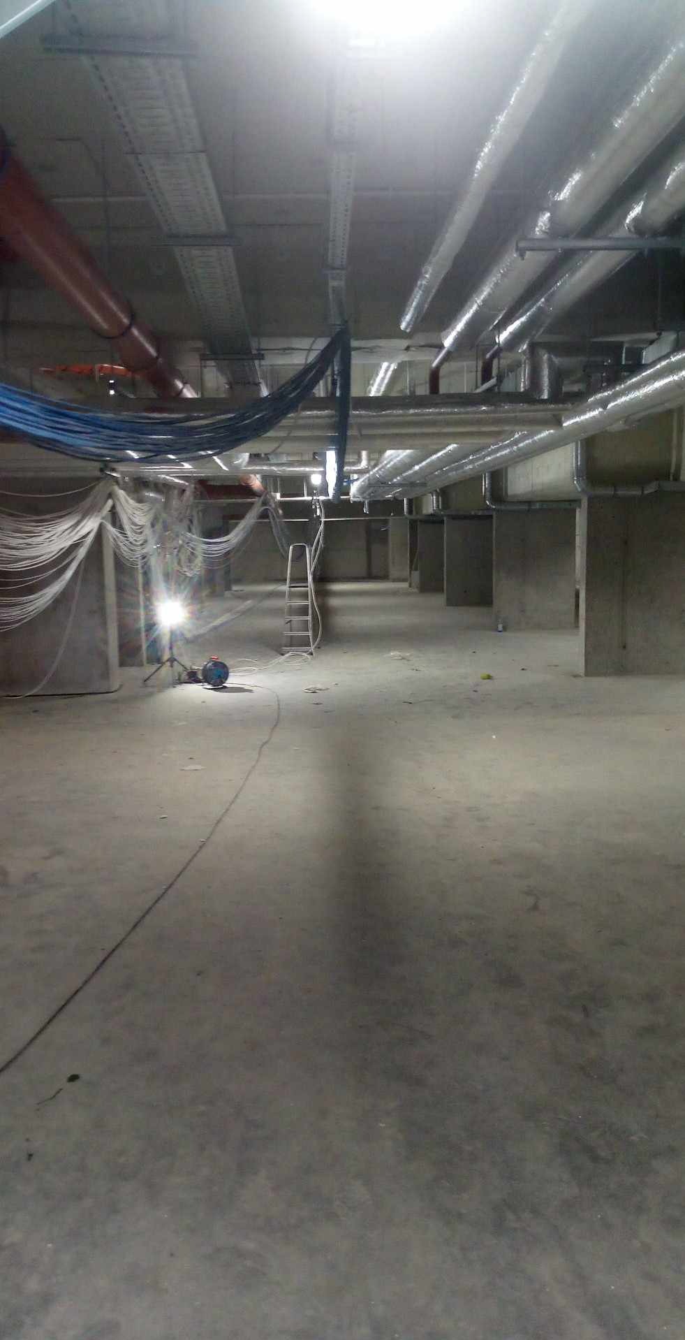 Wnętrze w stanie surowym z widocznymi instalacjami pod sufitem, przewody elektryczne zwisające ze ściany, drabina i lampa budowlana na statywie.