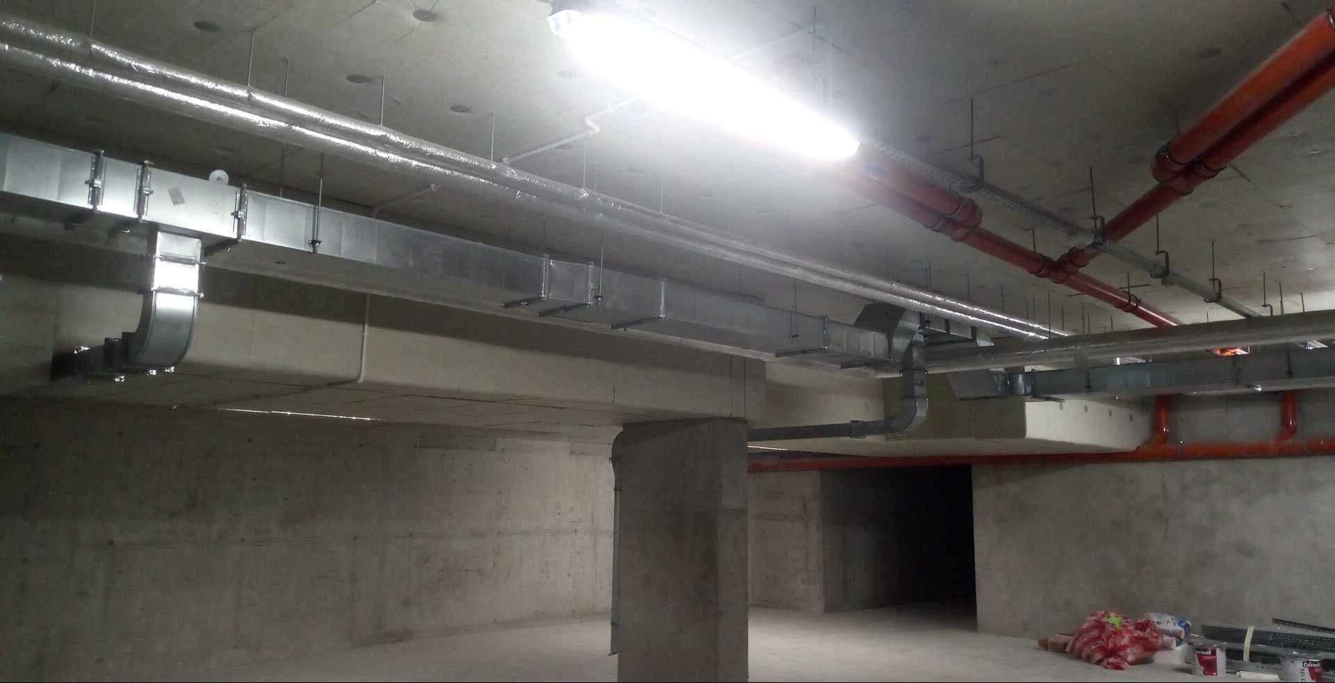 Surowy betonowy strop garażu z widocznymi instalacjami: srebrne kanały wentylacyjne, rury w kolorze ceglanej czerwieni, oświetlenie jarzeniowe. Na podłodze widoczne materiały budowlane i śmieci.