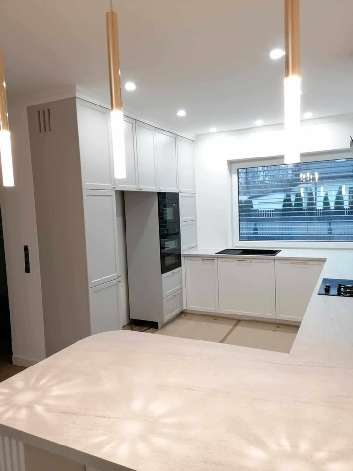 Jasna kuchnia na wymiar z białymi frontami, zabudowaną lodówką i piekarnikiem, oświetlona złotymi lampami wiszącymi, z widokiem na zewnątrz przez okno.