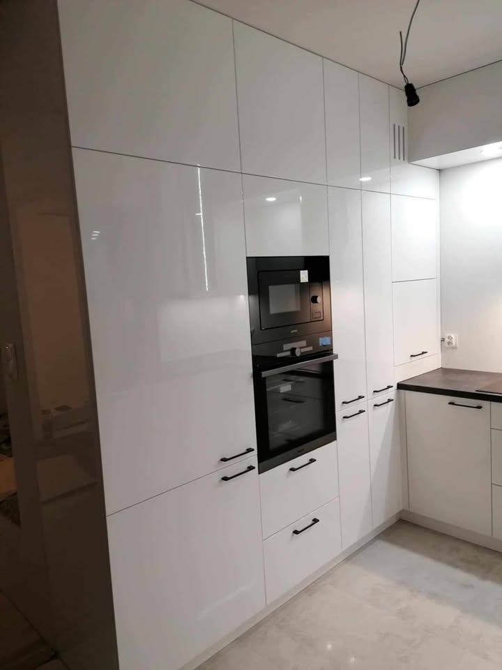 Biała zabudowa kuchenna z piekarnikiem i mikrofalówką, czarne, minimalistyczne uchwyty, błyszczące fronty, nowoczesny design, szara podłoga.