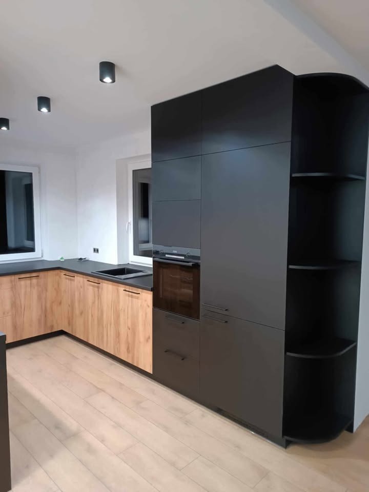 Nowoczesna kuchnia: drewnopodobne fronty szafek dolnych, czarne blaty i wysoka zabudowa z piekarnikiem oraz otwartymi półkami. Minimalistyczne oświetlenie punktowe.