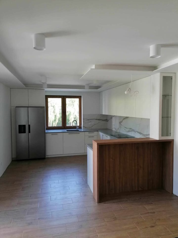 Nowoczesna kuchnia z białymi szafkami, marmurowym blatem i drewnianą wyspą. Widoczne oświetlenie punktowe i okno z widokiem na zieleń.