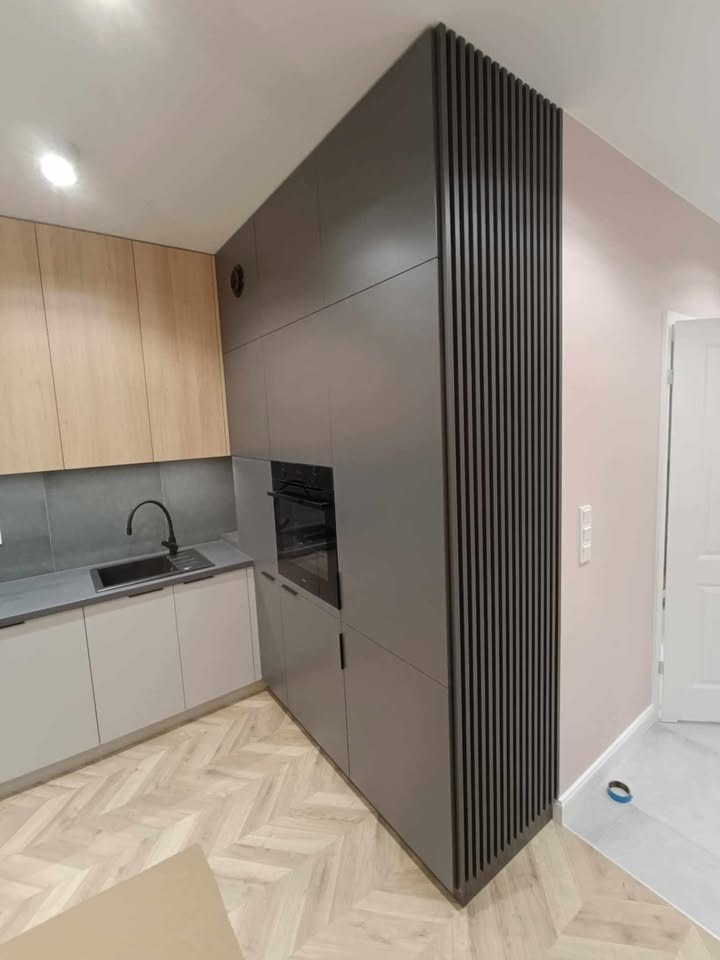 Szara zabudowa kuchenna z ryflowanym panelem bocznym, jasne drewniane fronty szafek wiszących, parkiet w jodełkę. Minimalistyczny design z czarną armaturą.