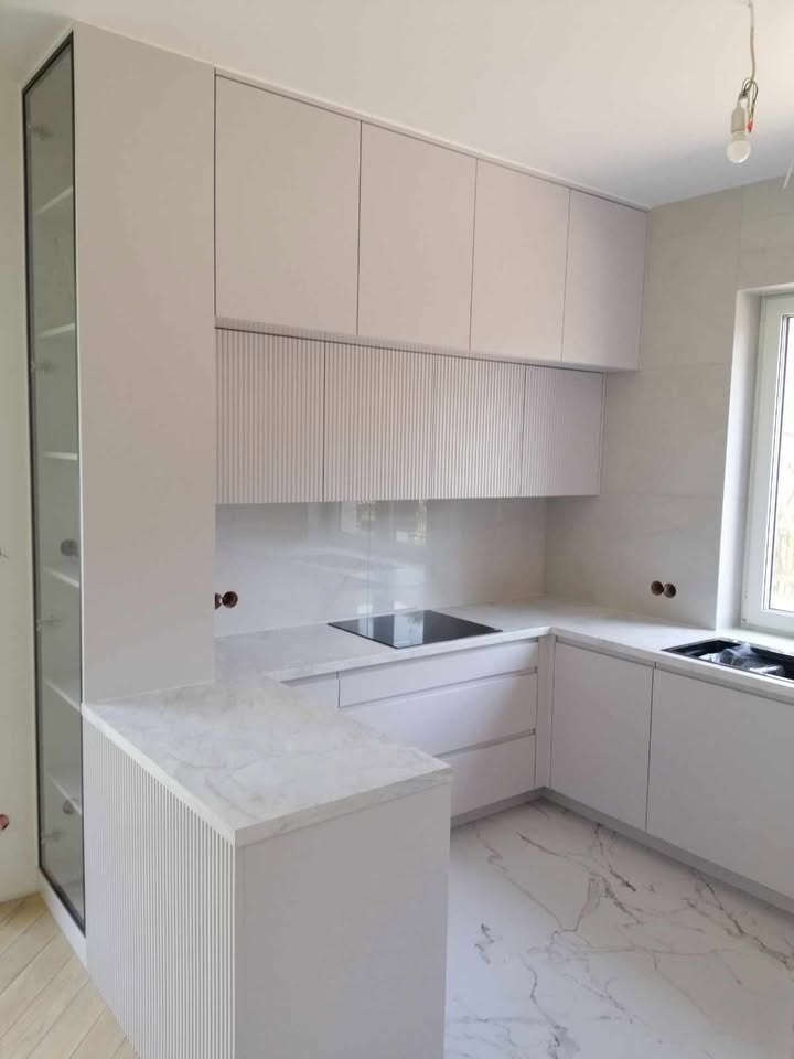 Nowoczesna, jasna kuchnia z białymi frontami, marmurowym blatem i podłogą. Ryflowane wykończenie szafek dodaje unikalnego charakteru. Minimalistyczny design.