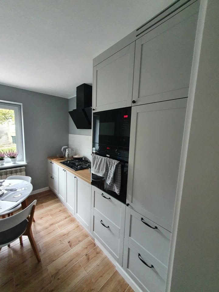 Jasna kuchnia z drewnianym blatem i czarnym okapem. Białe szafki z ciemnymi uchwytami. Widoczny stół z krzesłami i okno z kwiatami. Nowoczesny design.