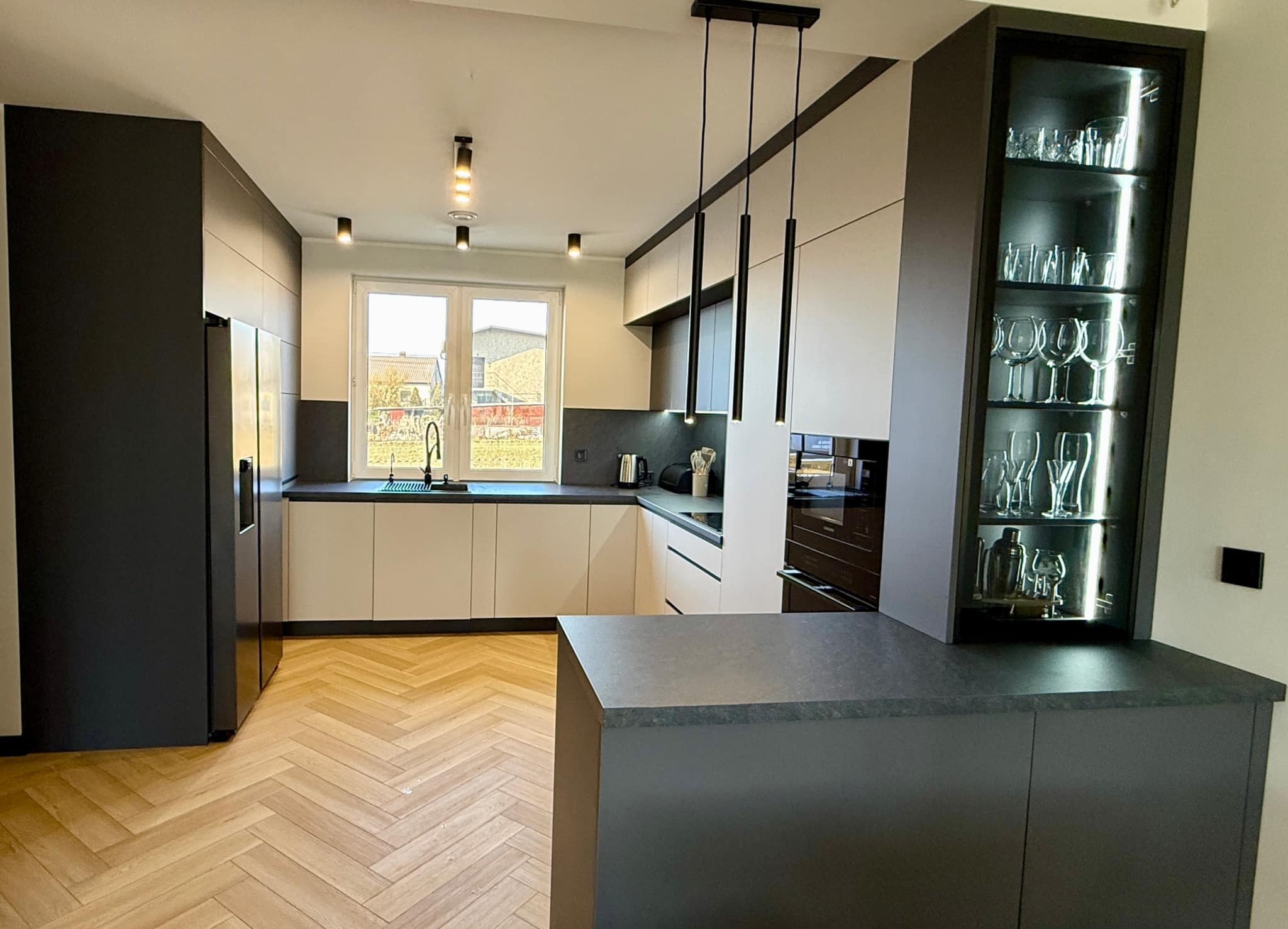 Nowoczesna kuchnia z wyspą, jasne szafki dolne, ciemne górne i wyspa. Parkiet w jodełkę i oświetlenie punktowe. Szklana witryna z oświetleniem LED.