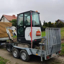 Biała minikoparka Bobcat na przyczepie z metalową kratą zabezpieczającą, widoczna na tle zieleni i zabudowań w pochmurny dzień.