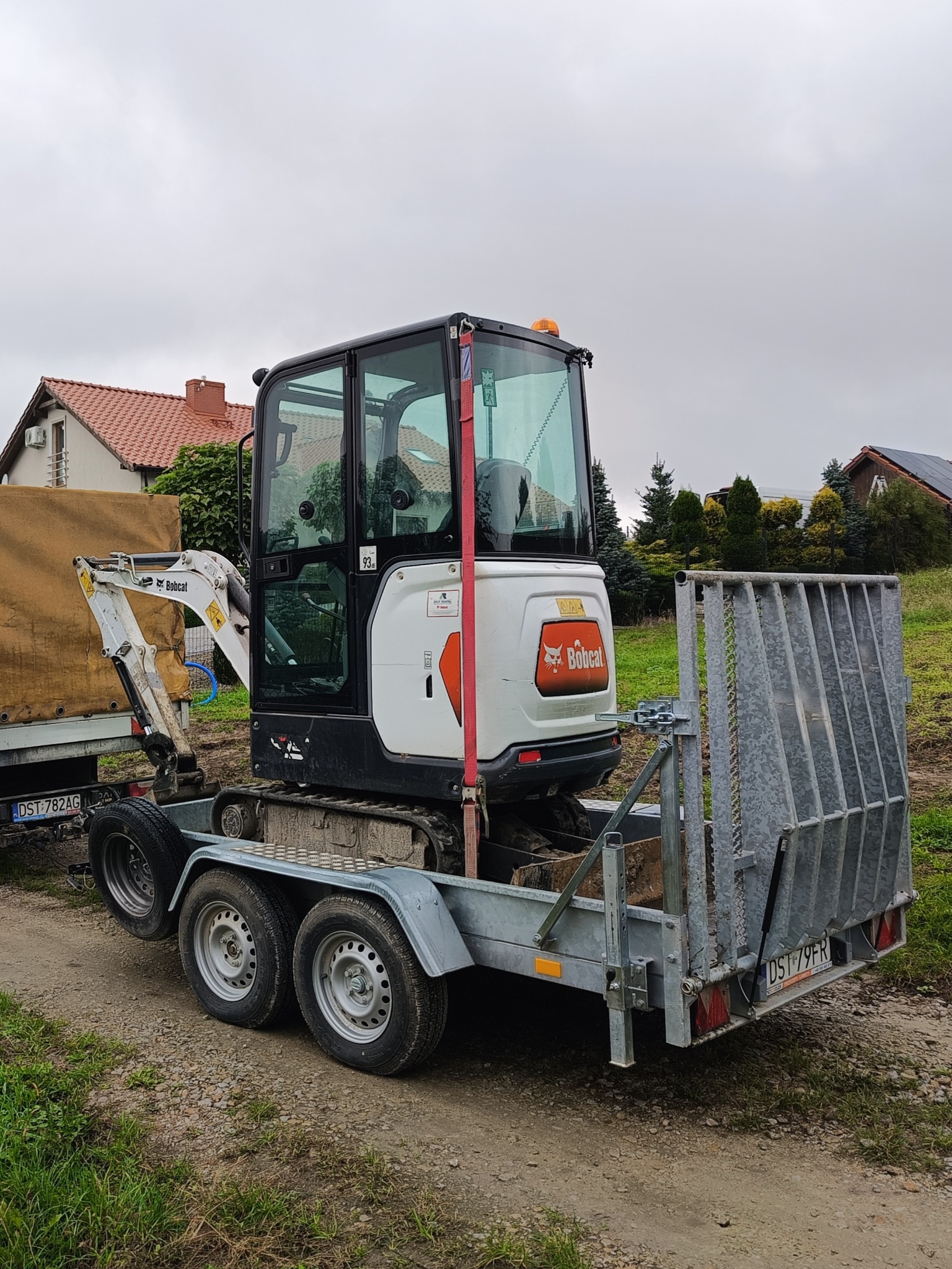 Biała minikoparka Bobcat na przyczepie z metalową kratą zabezpieczającą, widoczna na tle zieleni i zabudowań w pochmurny dzień.