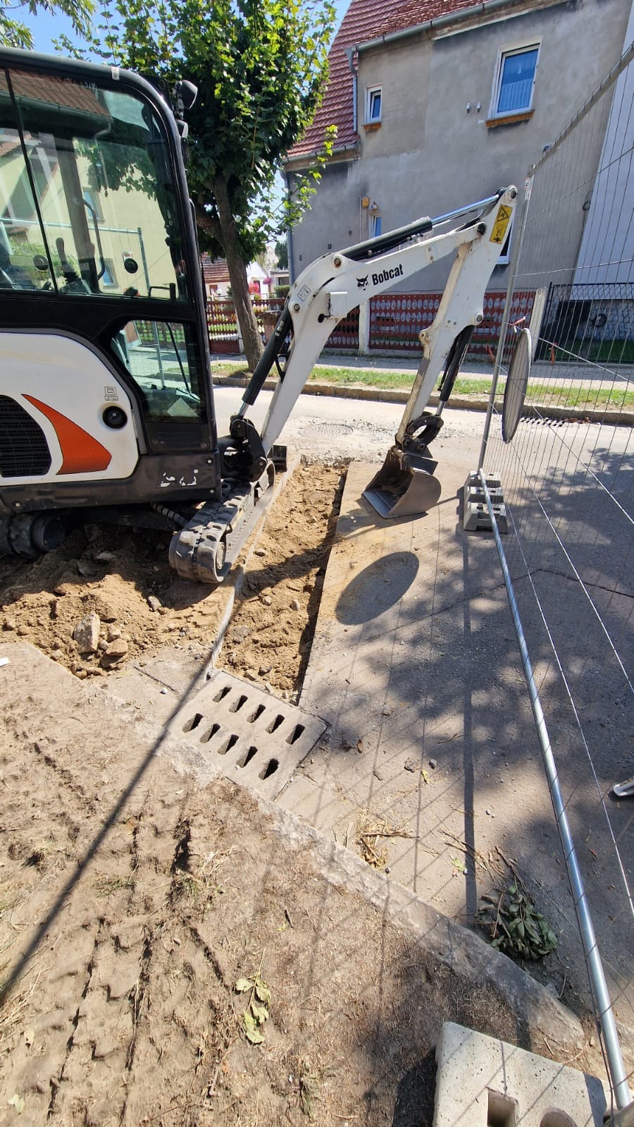 Biała minikoparka Bobcat wykonuje wąski wykop wzdłuż betonowego korytka odwadniającego na tle ulicy i zabudowań.