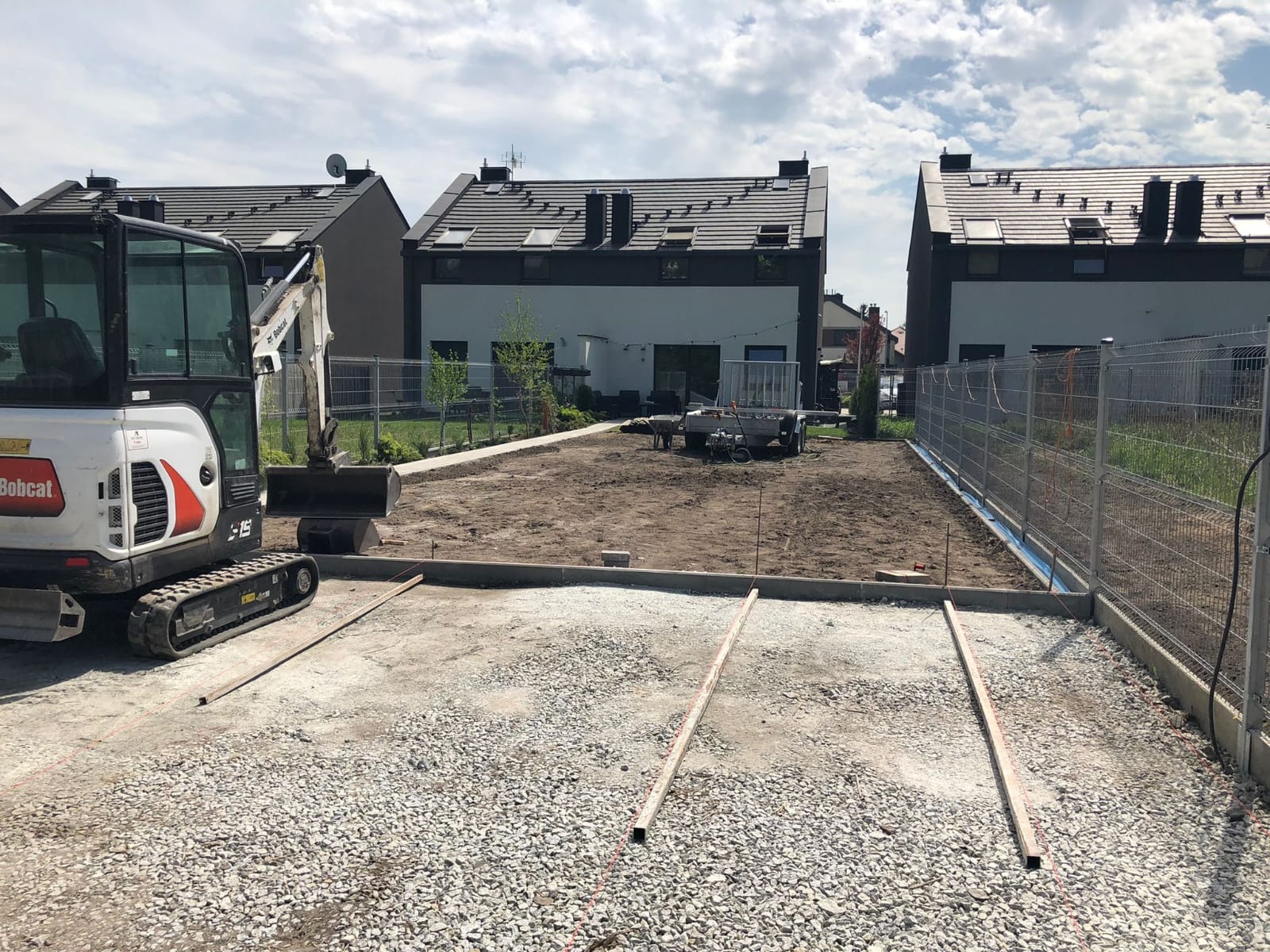 Przygotowany teren pod budowę z wysypanym kruszywem, widoczna minikoparka Bobcat i przyczepka, w tle zabudowa szeregowa.