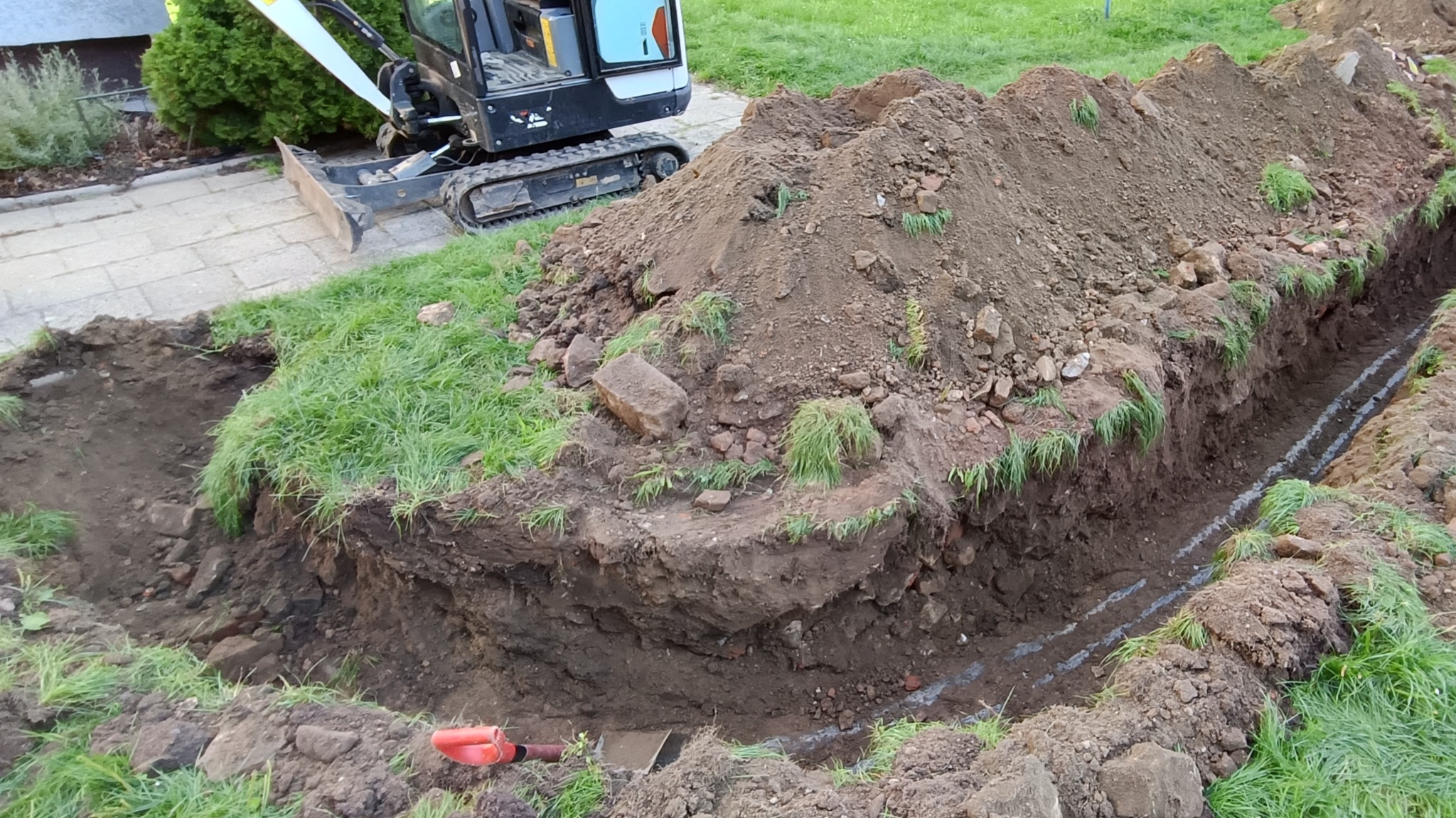 Wąski wykop w ziemi z widocznymi śladami po wyznaczeniu trasy, obok sterta ziemi i częściowo widoczna minikoparka na kostce brukowej.