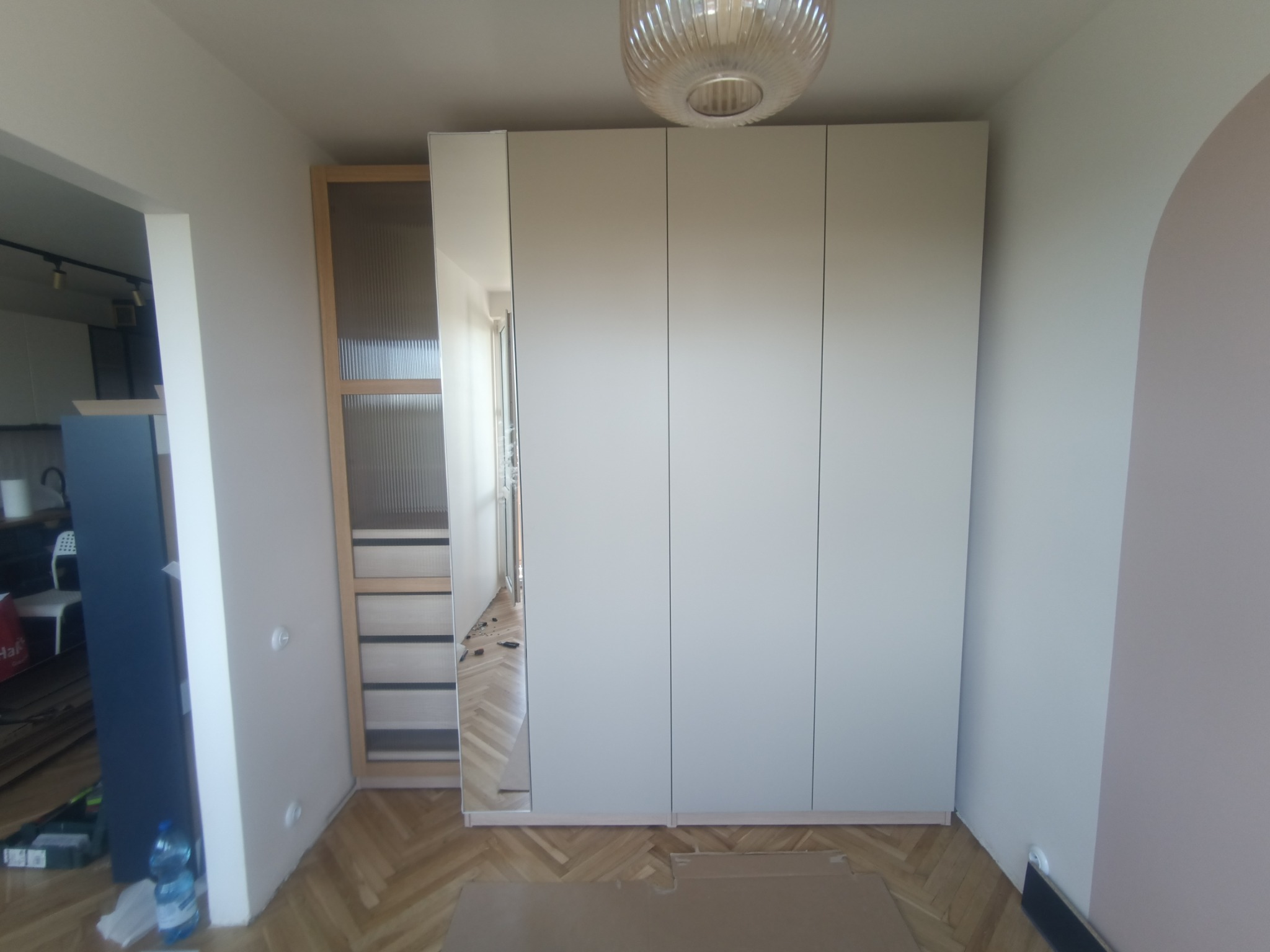 Nowoczesna szafa wnękowa z białymi frontami i lustrem, zamontowana w jasnym pomieszczeniu z parkietem, widoczny również element szafy z ryflowanym szkłem.